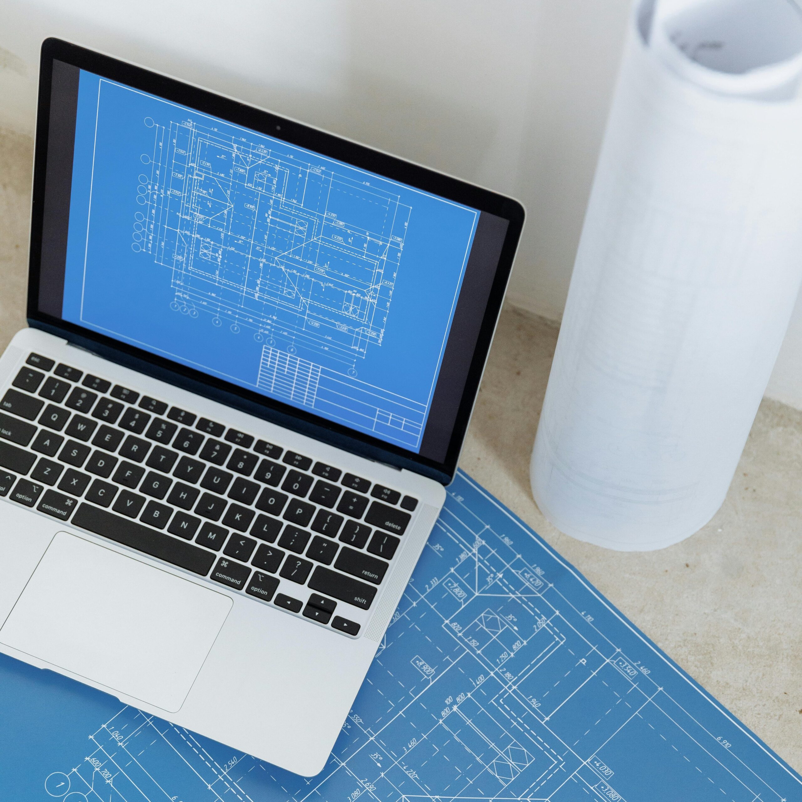 Laptop showing engineering blueprint with rolled paper and drawings on table.